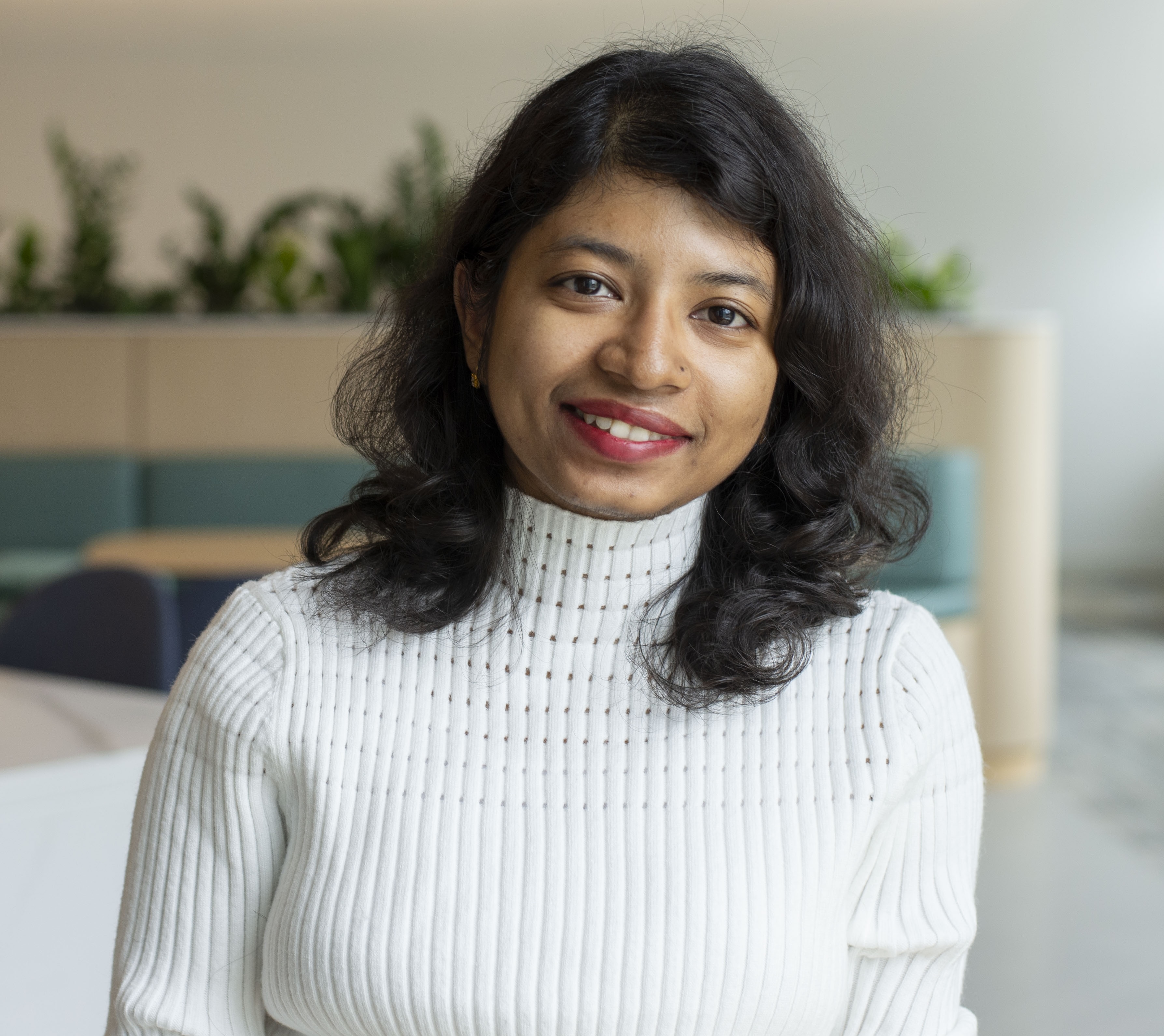 This image features a portrait of Rudaiba, a brown woman wearing a white ribbed turtleneck sweater. She has medium-length black hair with soft waves and a slightly parted style. The background is neutral.